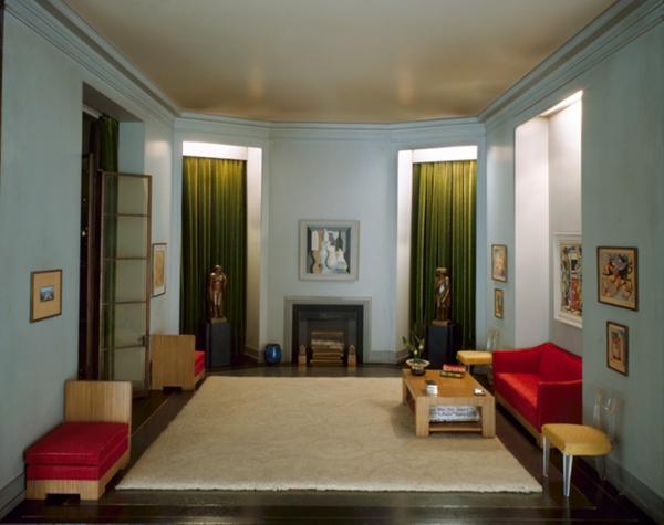 Miniature room with two tall windows framing a small Cubist painting and a fireplace, two golden scuptures set before the windows. At right, a small red couch is flanked by two Lucite chairs. At left, an open glass door is flanked by oak chaises with red cushions. A plush cream rug fills the space.